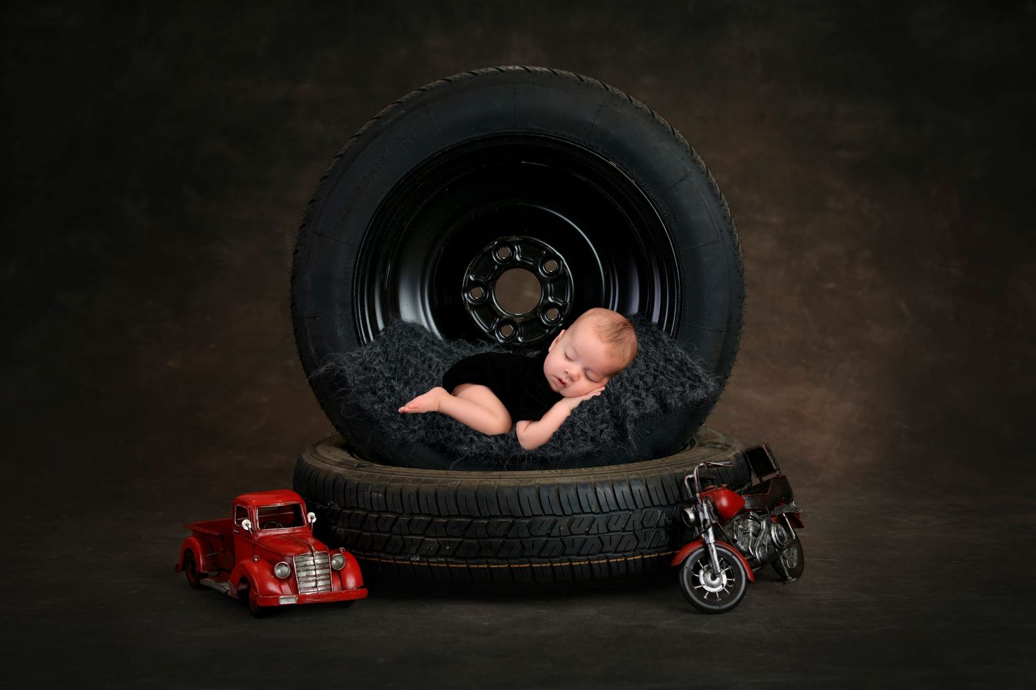 Newborn baby sleeping inside a large tire with vintage toy cars nearby.