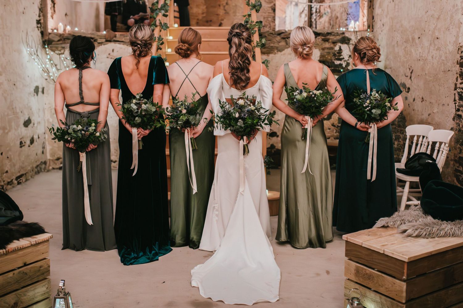 Row of bridesmaids in mix of dark and light dresses holding greenery bouquets in rustic venue.
