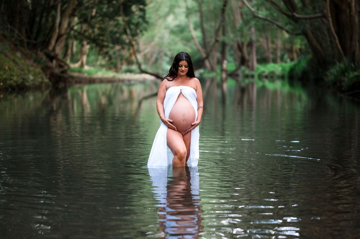 Artistic maternity photo of woman in white fabric standing in tranquil forest stream with reflective water.