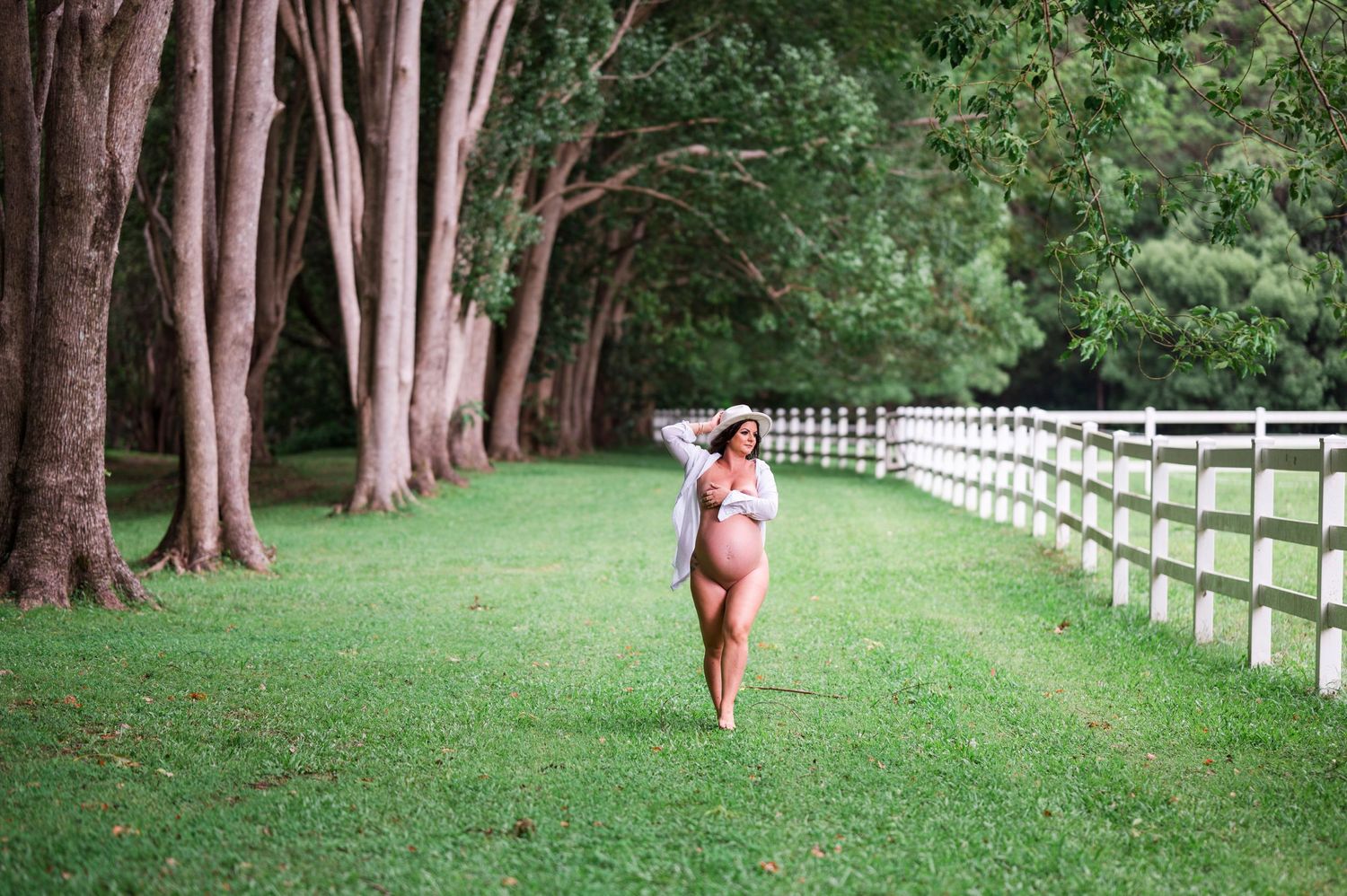 Expectant mother walking along tree-lined path with white fence in ethereal outdoor setting.