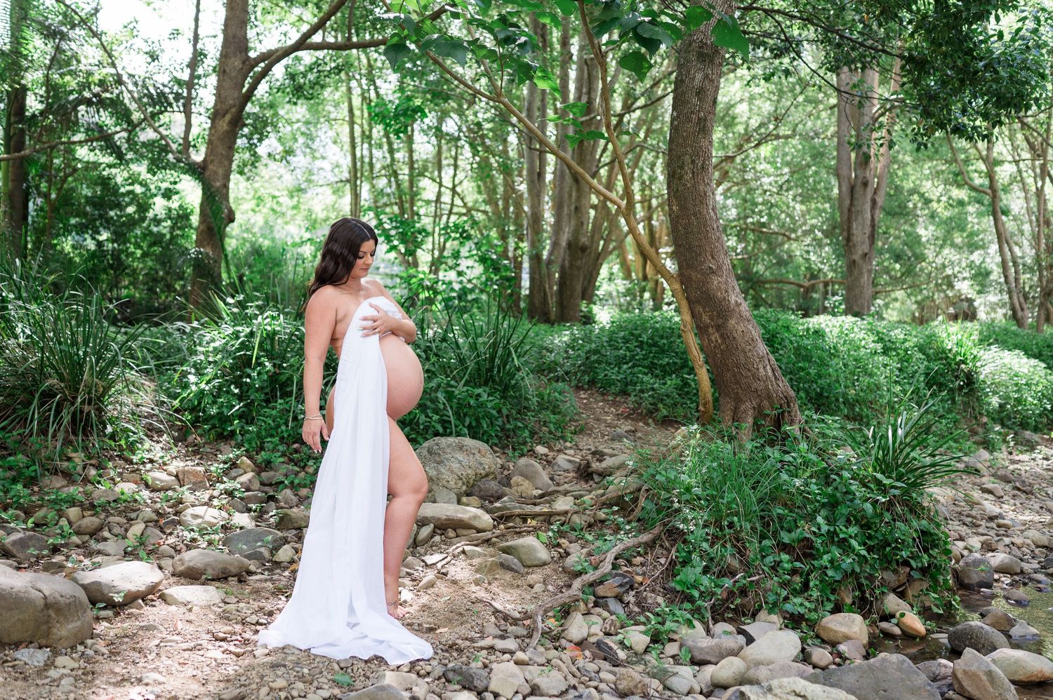 A woman in a flowing white dress poses elegantly in a lush forest setting.