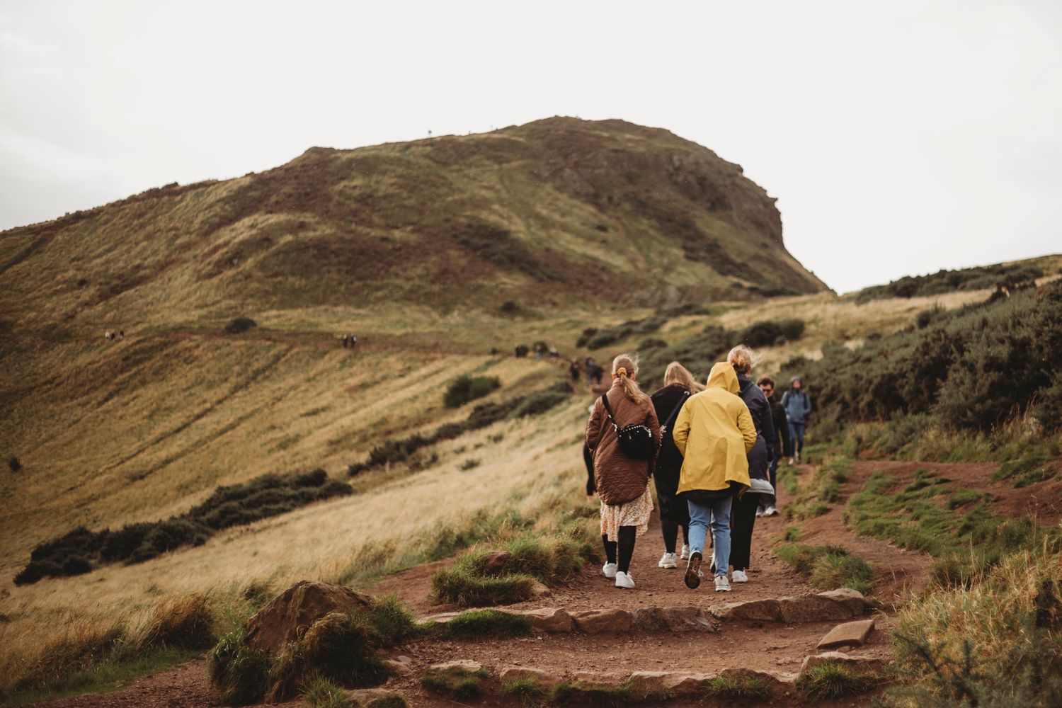 Hikers walking up dirt trail on steep grassy hillside under cloudy sky.