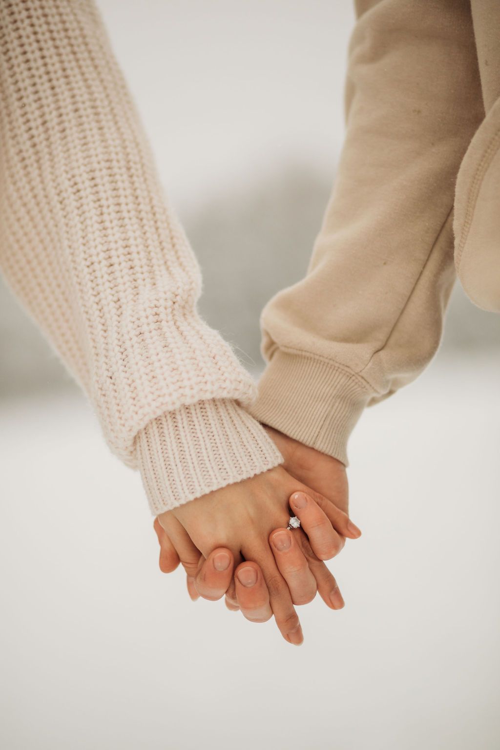 Close-up of intertwined hands showing engagement ring against snowy background.