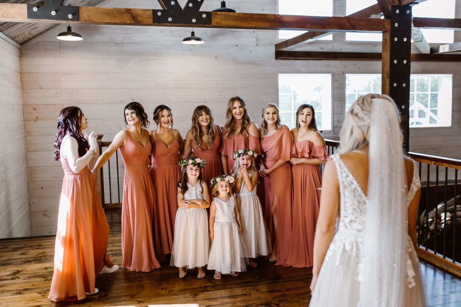 Wedding party in terracotta colored dresses admiring white bridal gown hanging in rustic venue.