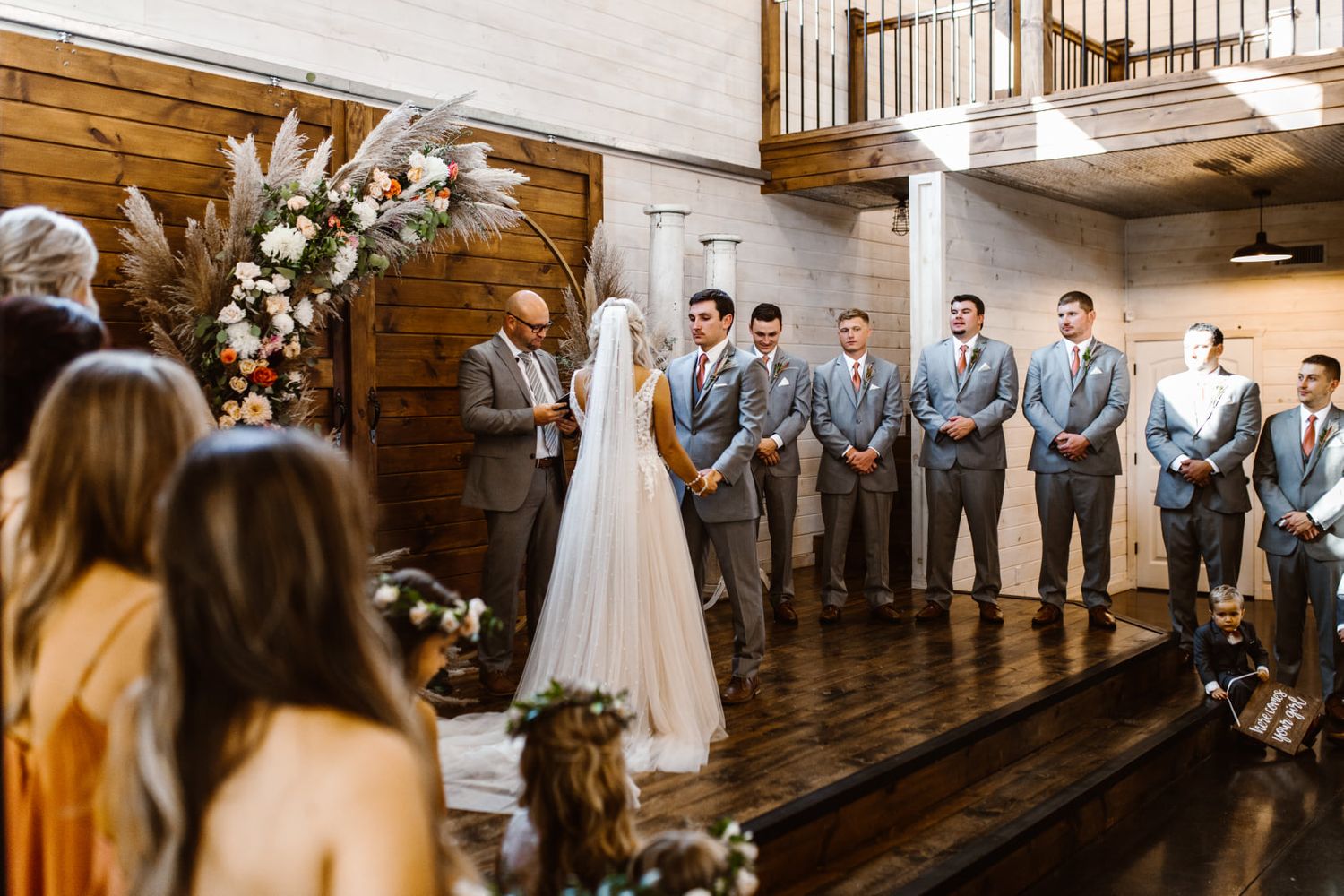Wedding ceremony in rustic wooden venue with floral arch and grey suited groomsmen.