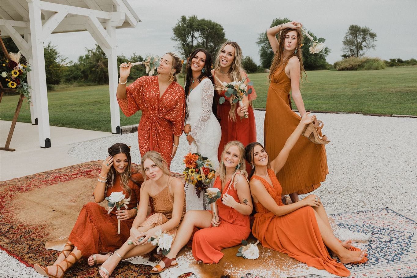 Bridal party in rust and orange dresses poses together outdoors under white pergola.