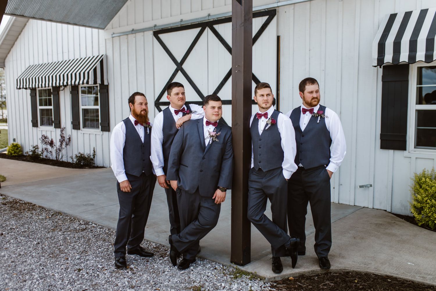 Groomsmen in matching navy vests pose casually outside a white barn-style venue.