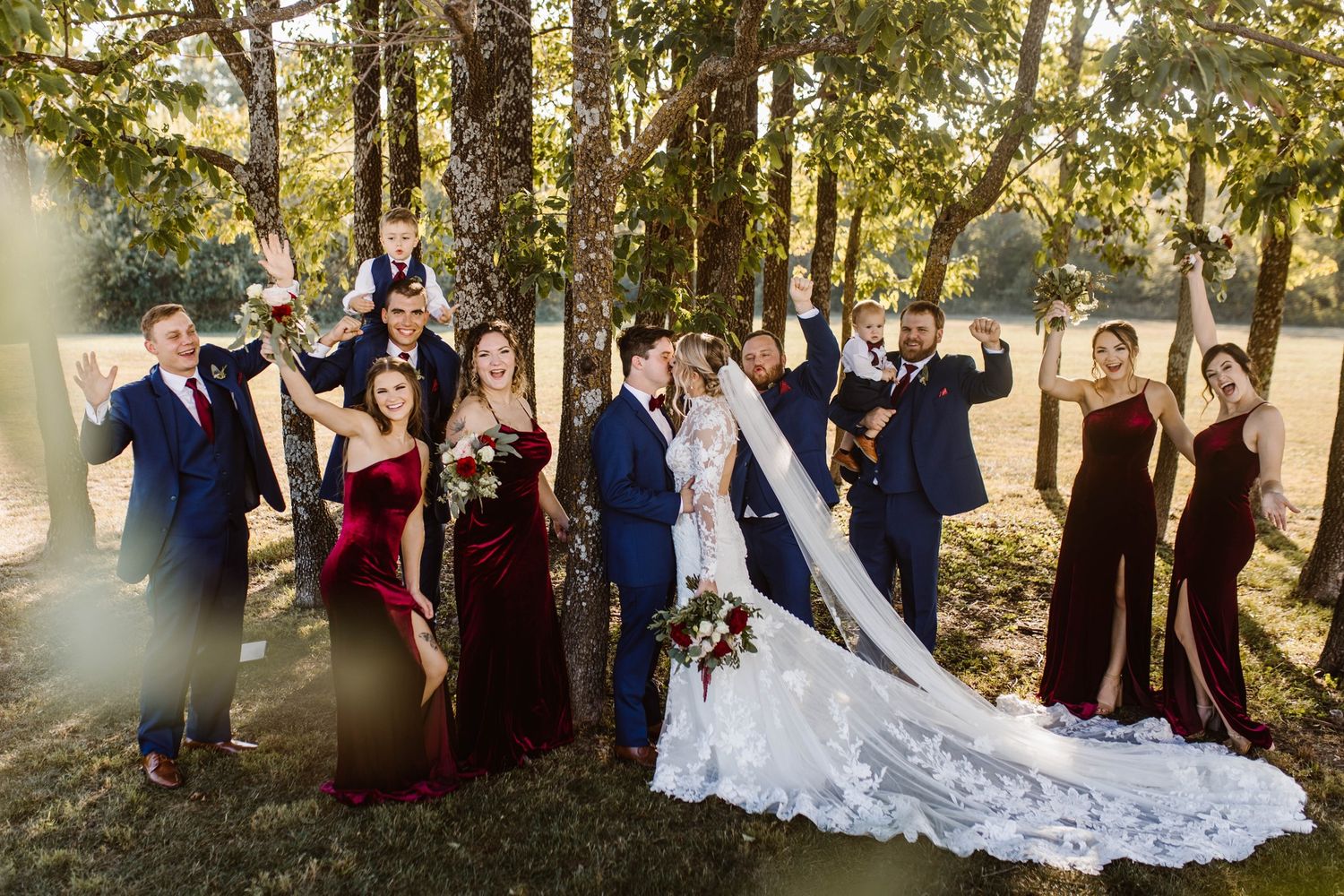 Wedding party poses in navy and burgundy attire against a forest backdrop on a sunny afternoon.