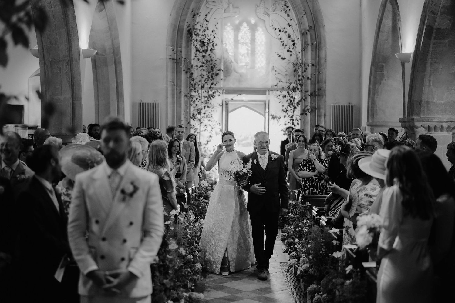 bride wiping a tear as she walks down the church aisle with her dad to meet her groom at st Nicholas church in Oxfordshire