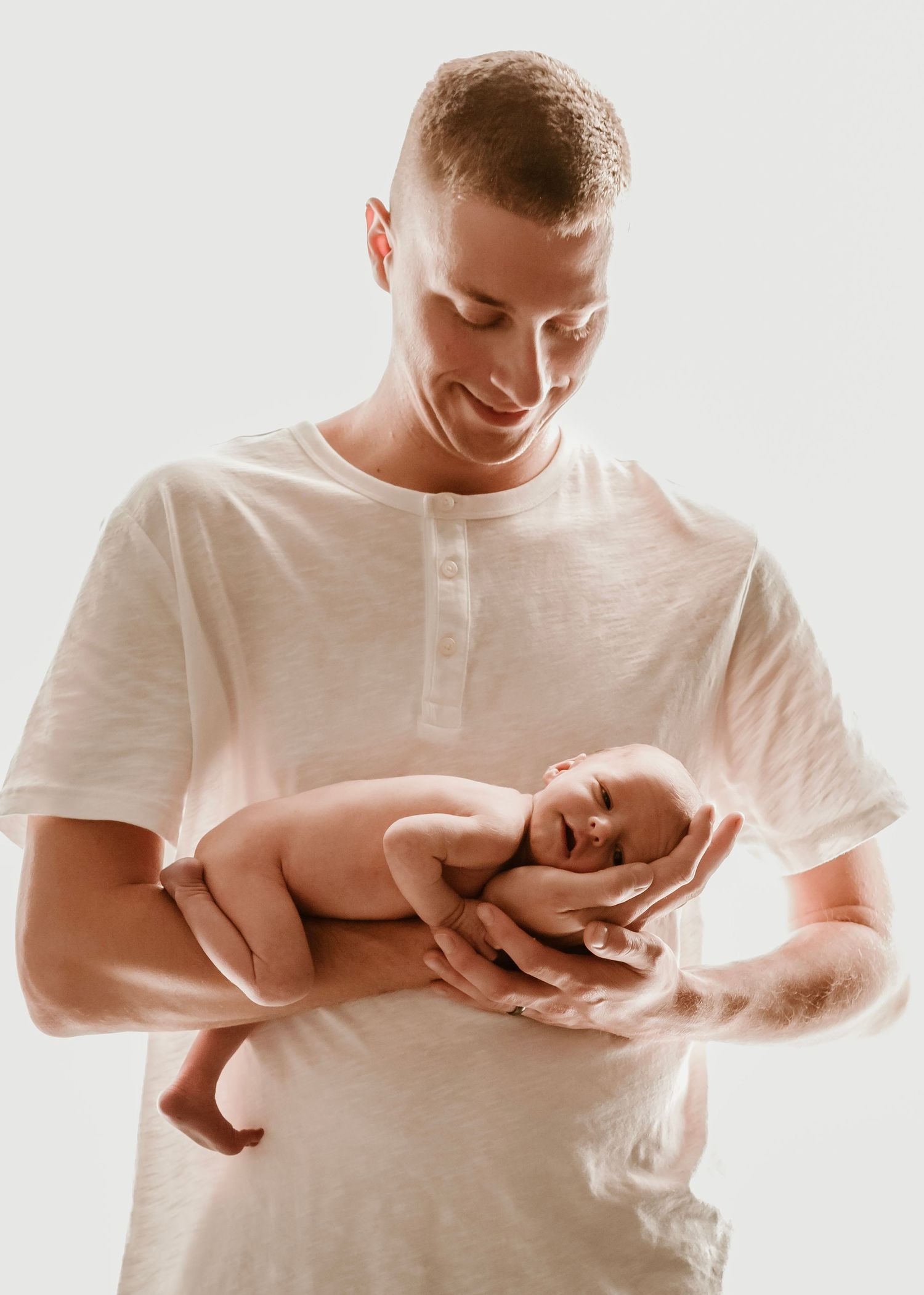 Parent in white shirt tenderly cradles naked newborn baby against bright background.