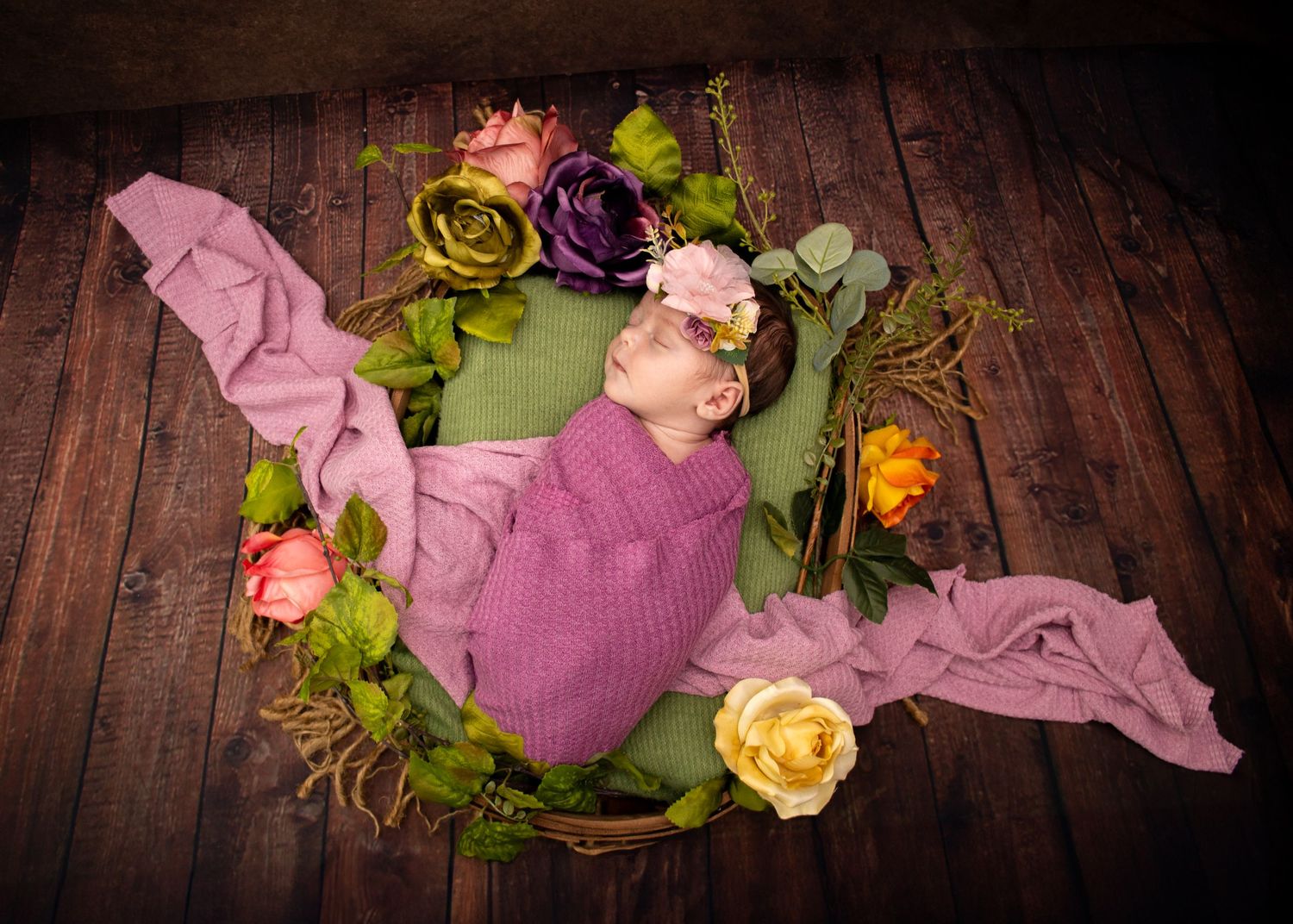 Newborn in pink wrap surrounded by colorful roses on wooden floor.