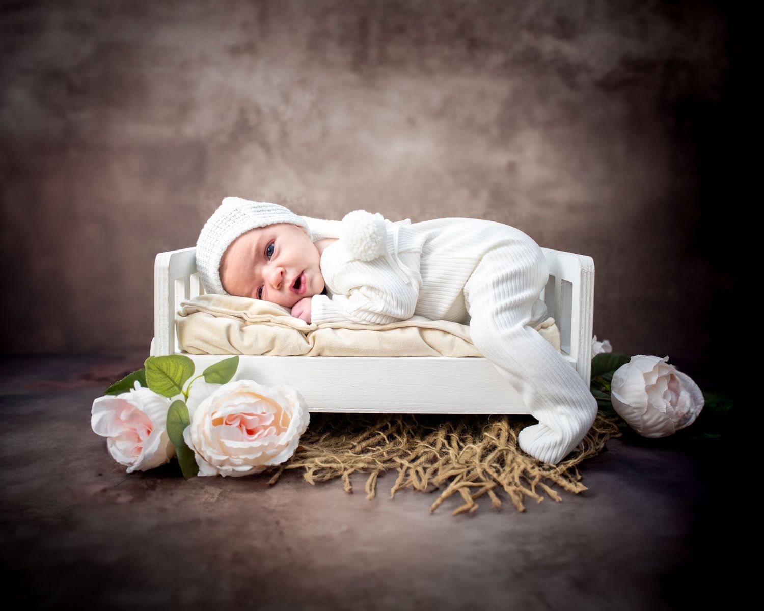 Newborn baby sleeping peacefully on white miniature bed with pink roses in a moody studio setup.