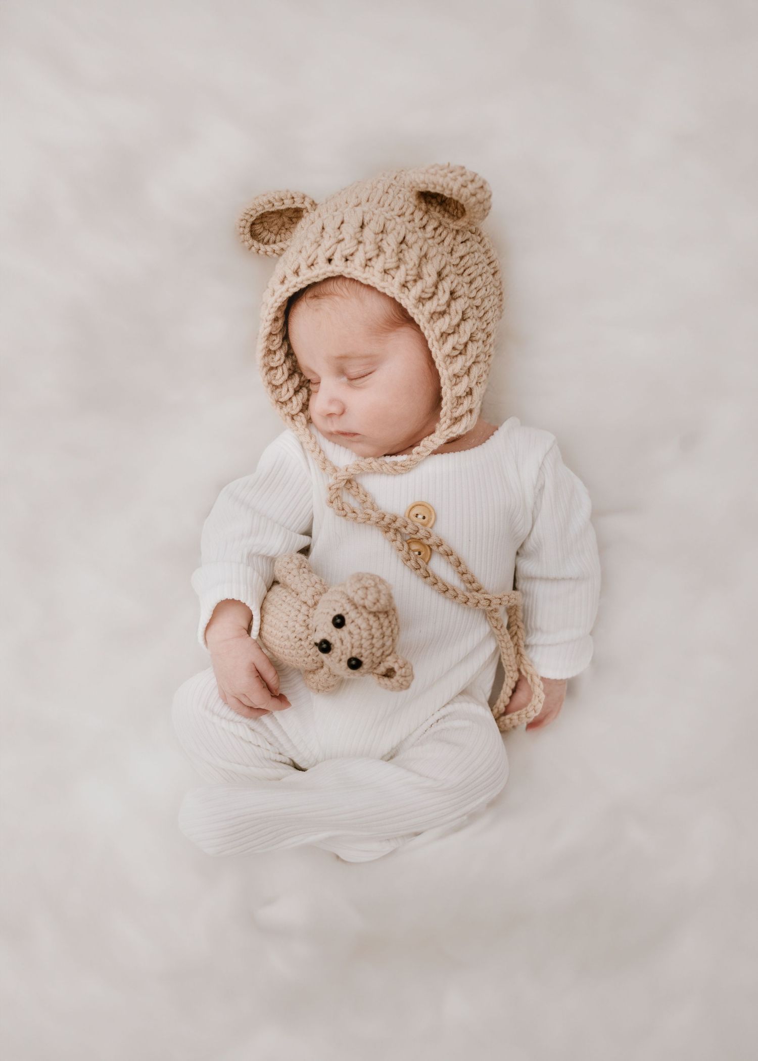 Sleeping baby wearing bear hat and holding teddy bear on white background.