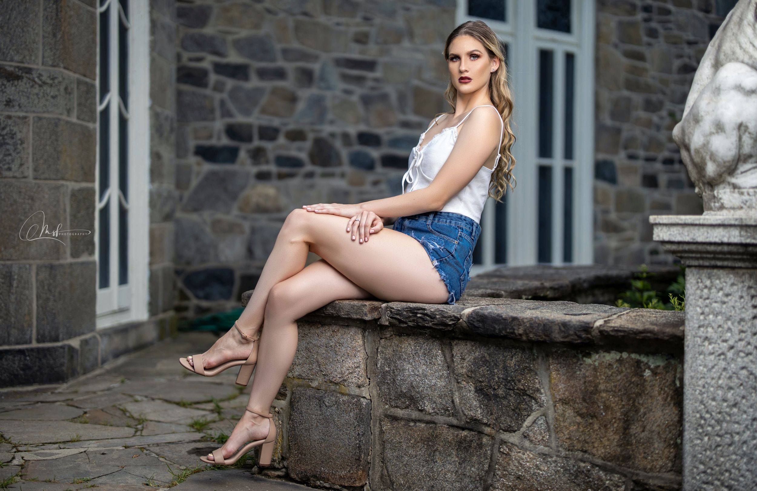 Woman in white top and denim shorts sits casually on stone wall near historic building.