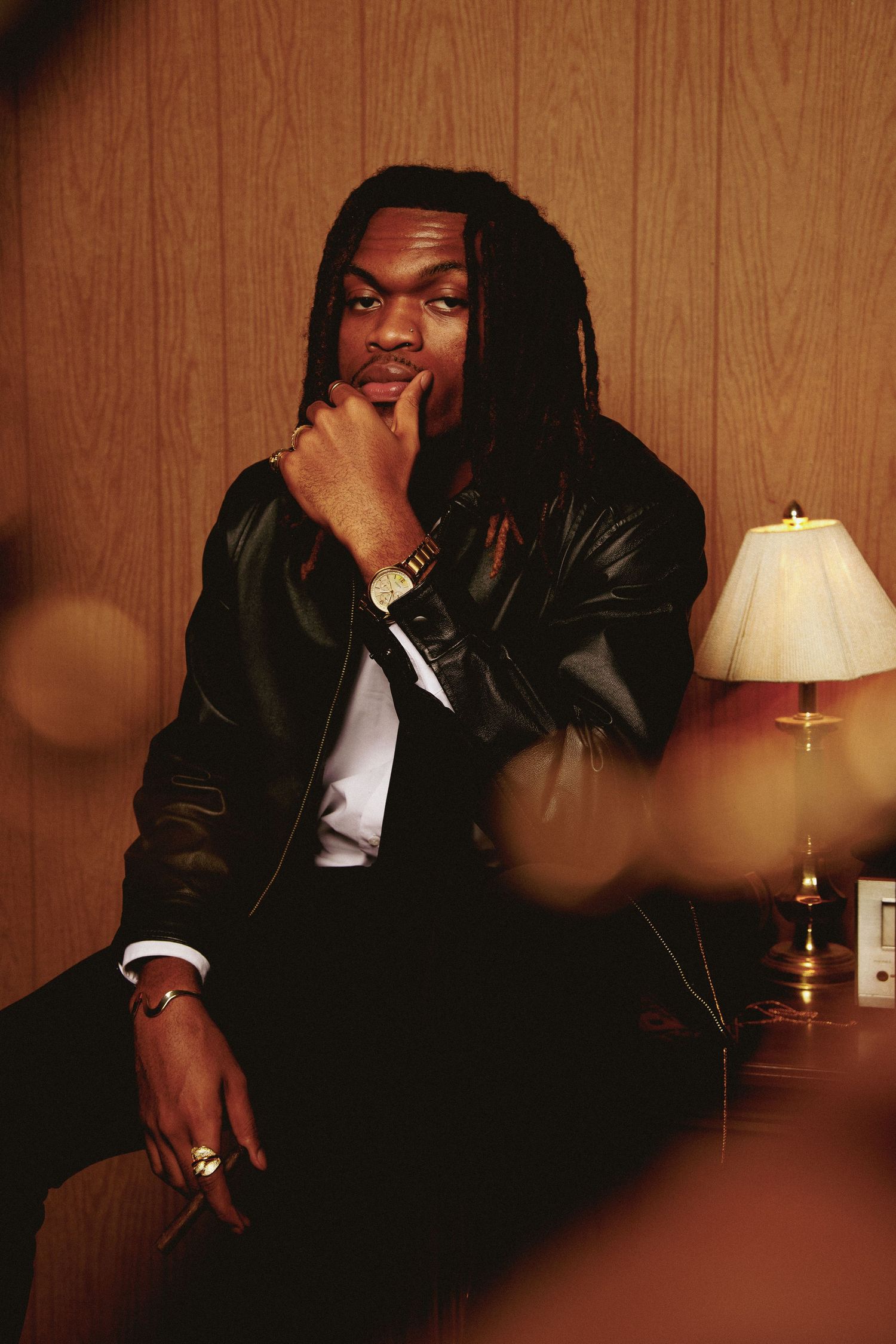 Stylish figure in leather jacket poses thoughtfully against wood paneling with table lamp in background.