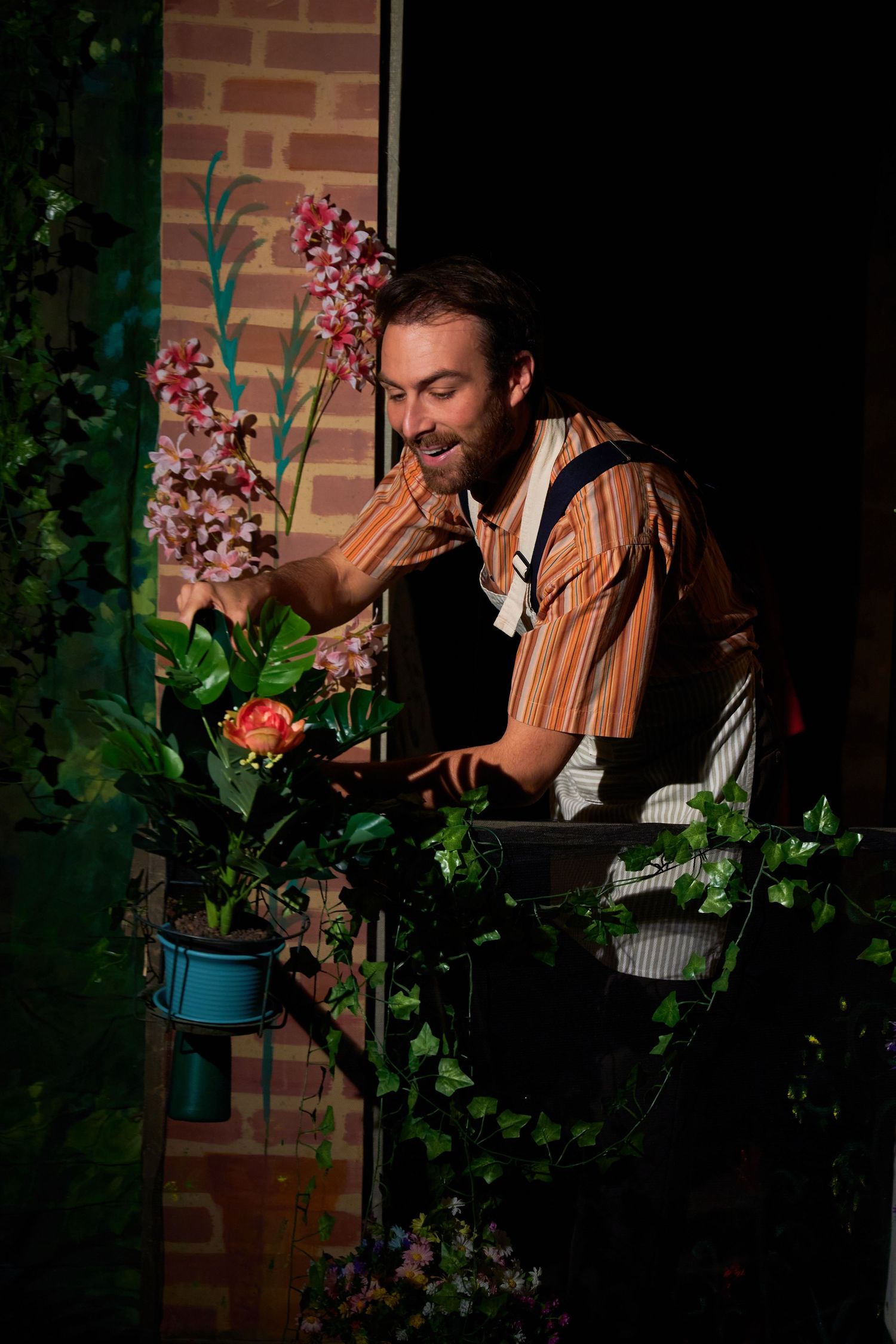 A performer admires flowers in a garden scene with brick backdrop and theatrical lighting.