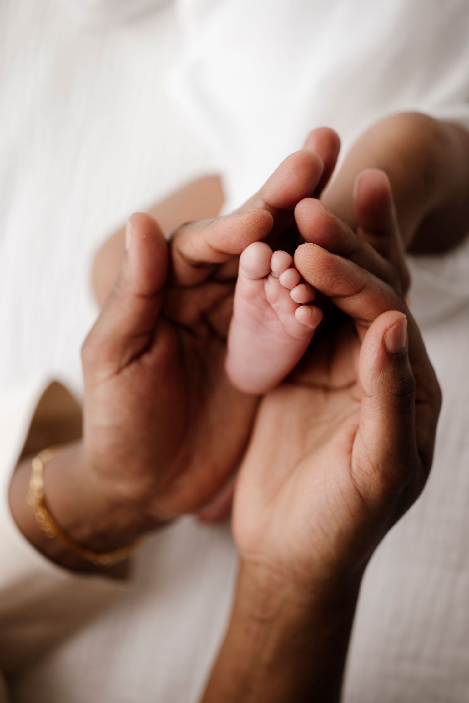 Gentle hands cradle tiny newborn feet against soft white fabric in intimate close-up.