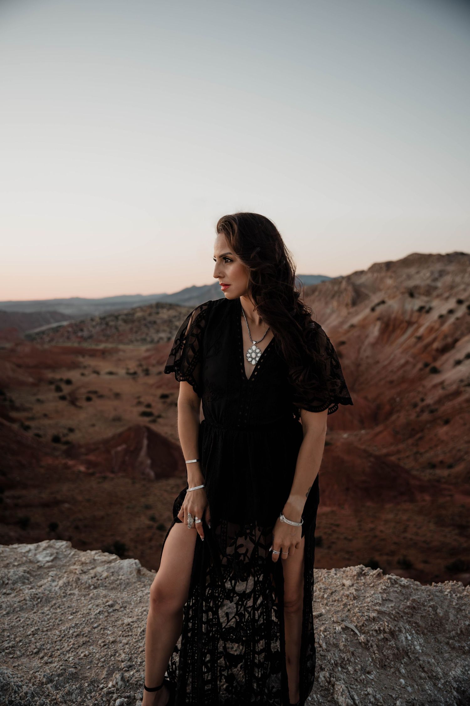 Woman in black dress sits on rocky outcrop overlooking desert landscape at sunset.