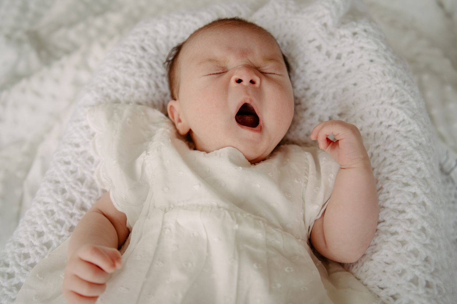 Baby in white dress yawning while lying on lace blanket.