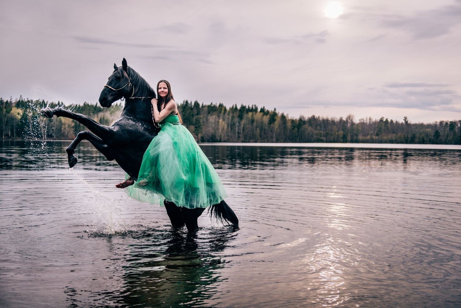 Woman in green gown riding black horse rearing up in lake with forest backdrop.