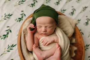 Sleeping newborn in green bonnet resting in wooden bowl with leafy backdrop.
