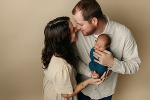 Intimate newborn family portrait with parents cradling tiny baby against neutral background.