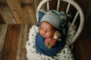 Sleeping newborn wearing knit hat and wrap in white wooden chair setting.