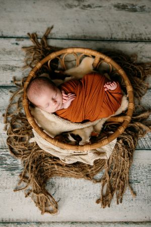 Newborn in orange wrap sleeping peacefully in rustic woven basket on wooden floor.