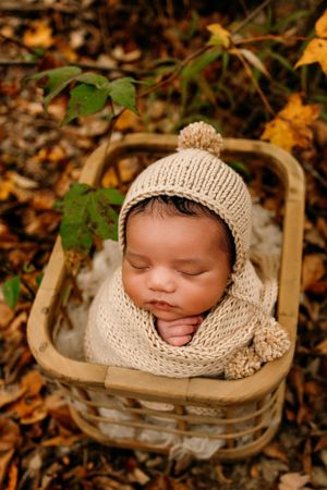 Sleeping baby in knit bonnet nestled in wooden basket surrounded by autumn leaves.