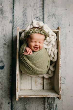 Baby wrapped in sage green fabric sleeping in rustic wooden box against distressed background.