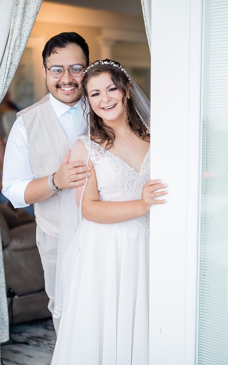 Wedding couple in white attire sharing joyful moments by a window with sheer curtains.