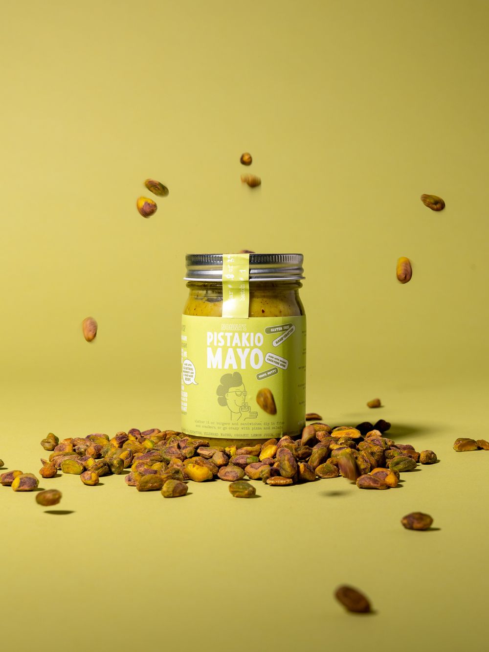 Artistic product shot of pistachio mayo jar with floating nuts on yellow background.