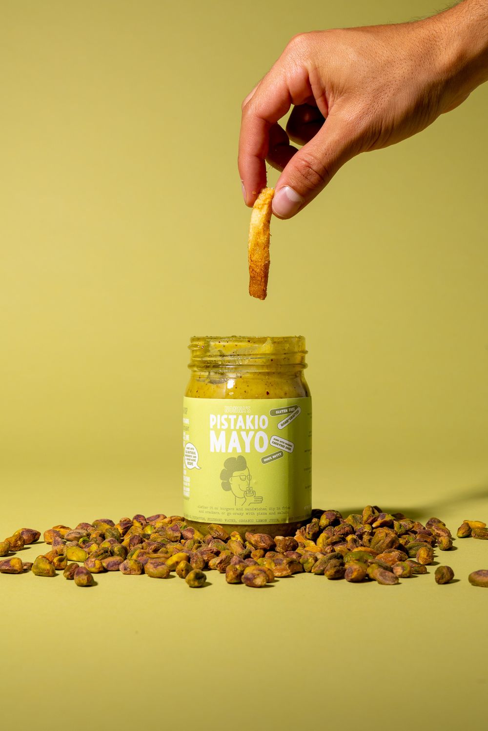 Hand dipping food into a jar of pistachio mayo surrounded by nuts on yellow background.