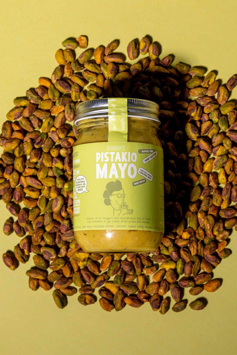 Jar of pistachio mayo surrounded by scattered pistachios on yellow background.