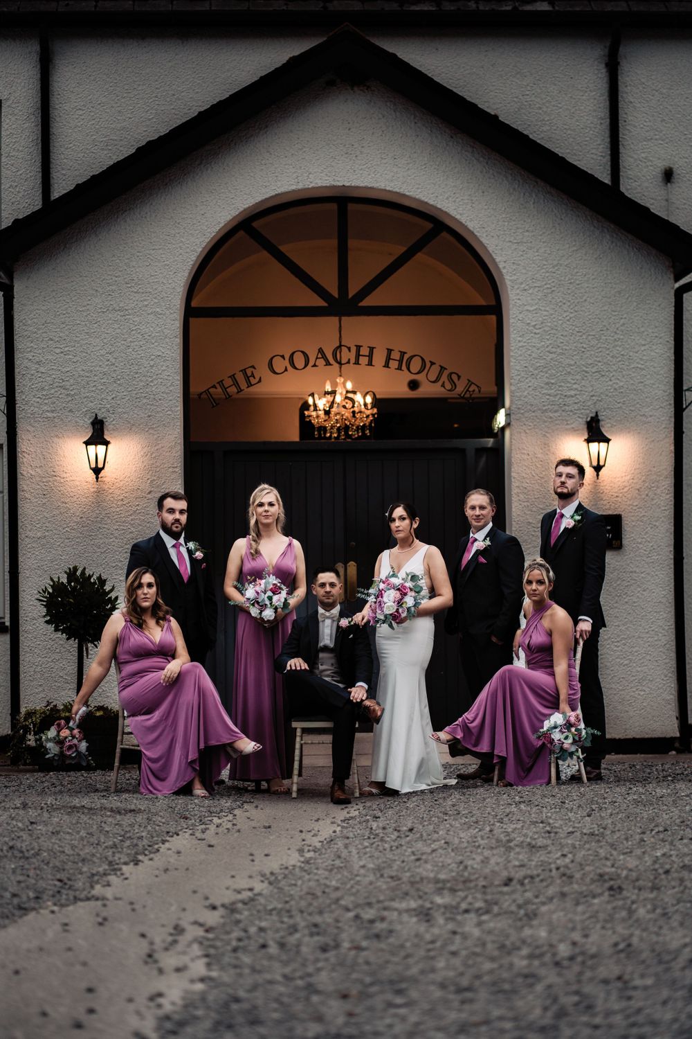 Wedding party posed in front of The Coach House entrance with lit lanterns at dusk.