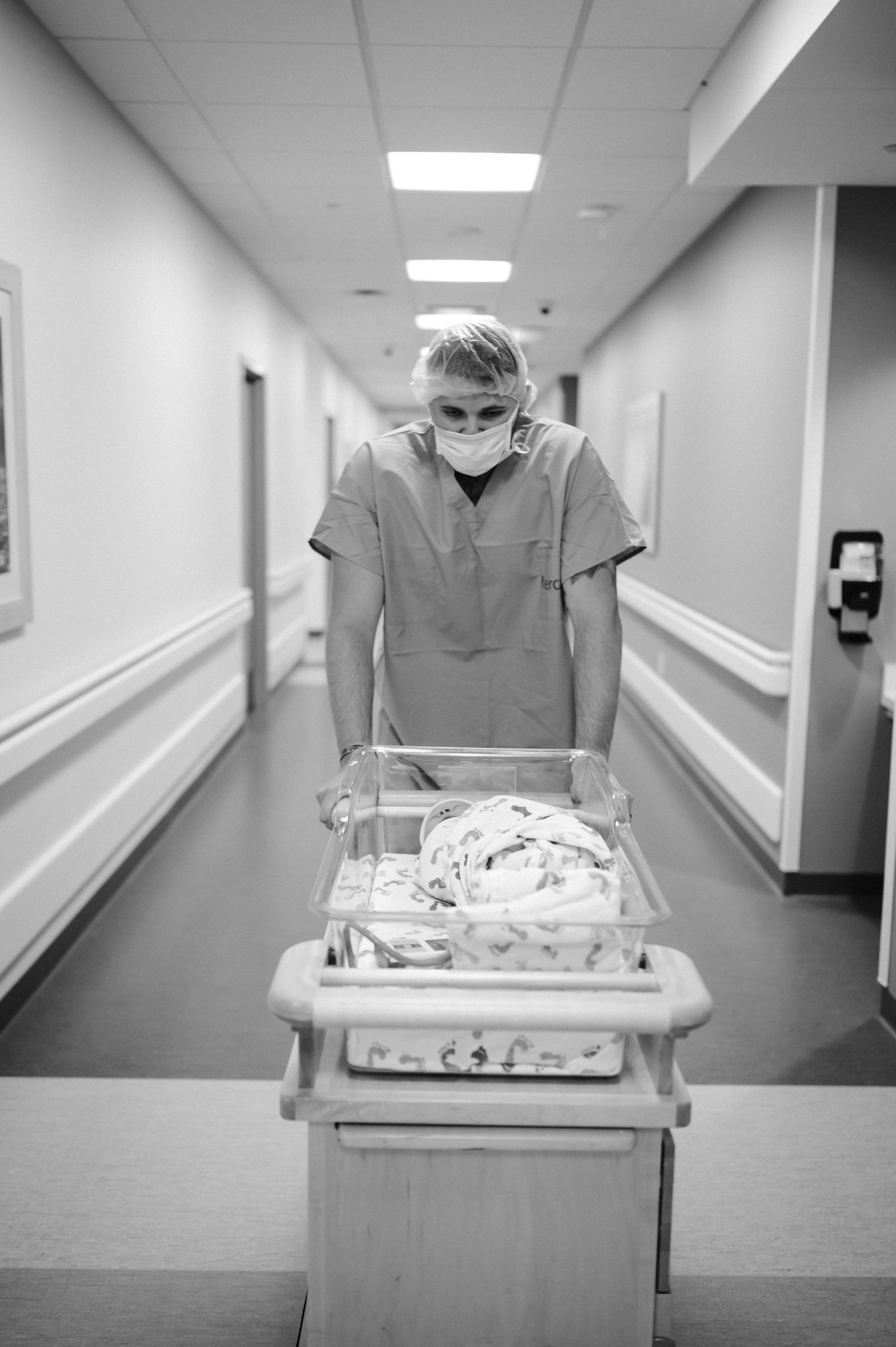 Healthcare worker in PPE pushing medical cart down hospital hallway in black and white.