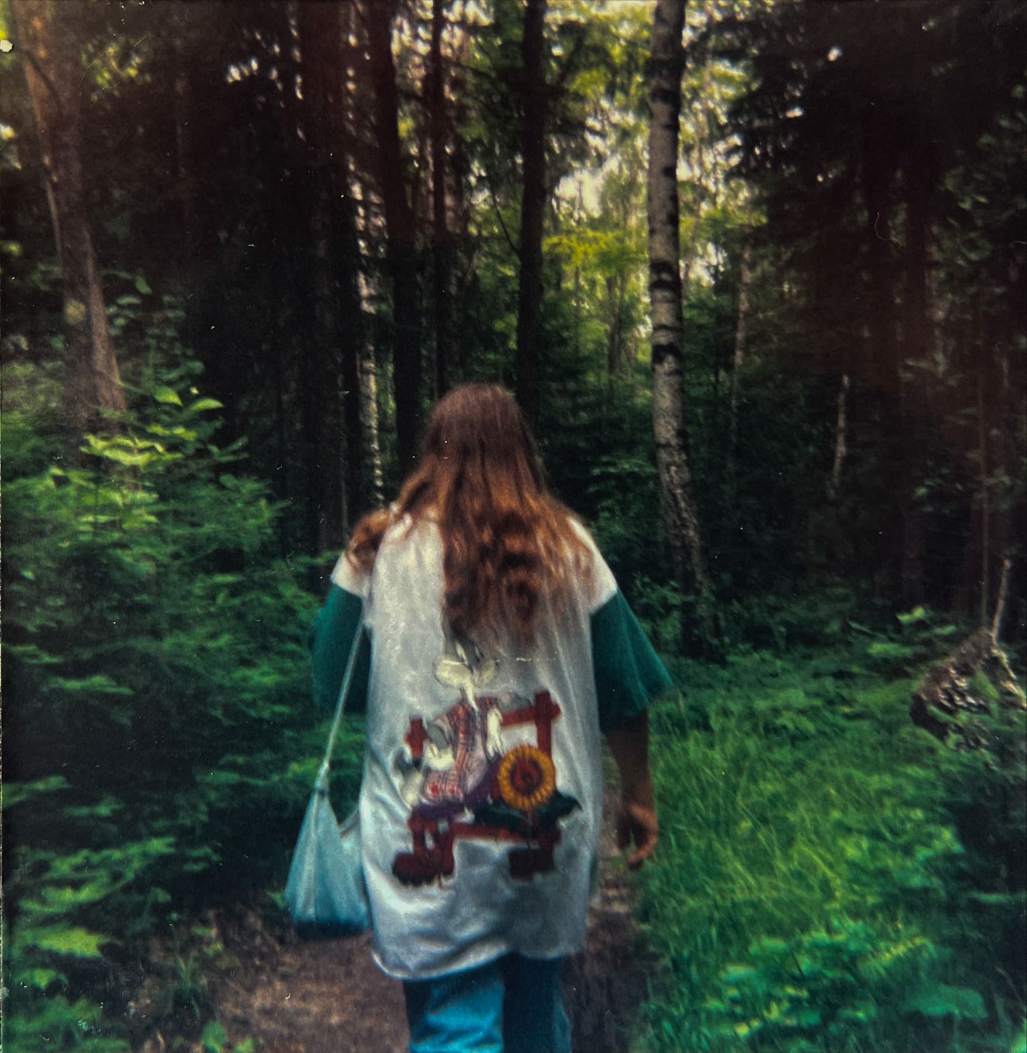 Retro square photograph of woman's back while walking through a wooded area. Taken on film.