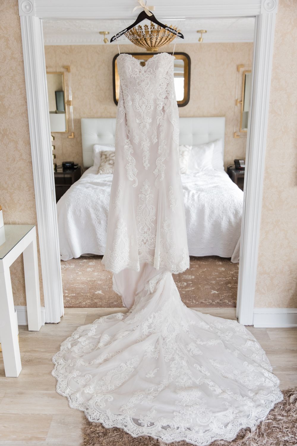 White lace wedding dress with long train hanging on display in bedroom.