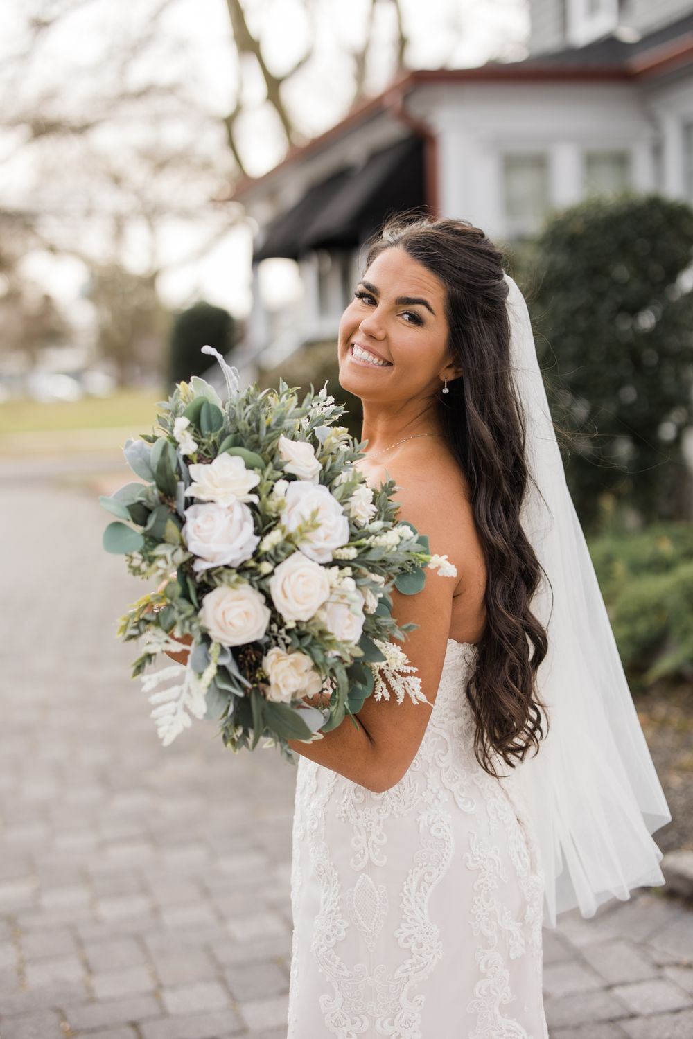 A bride in a lace gown holds a white and green bouquet while standing outdoors.