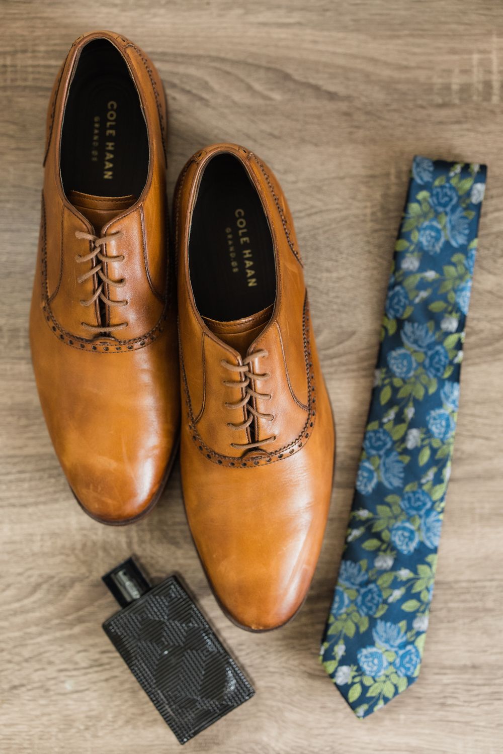 Brown leather dress shoes with floral tie and cologne bottle arranged on wooden surface.