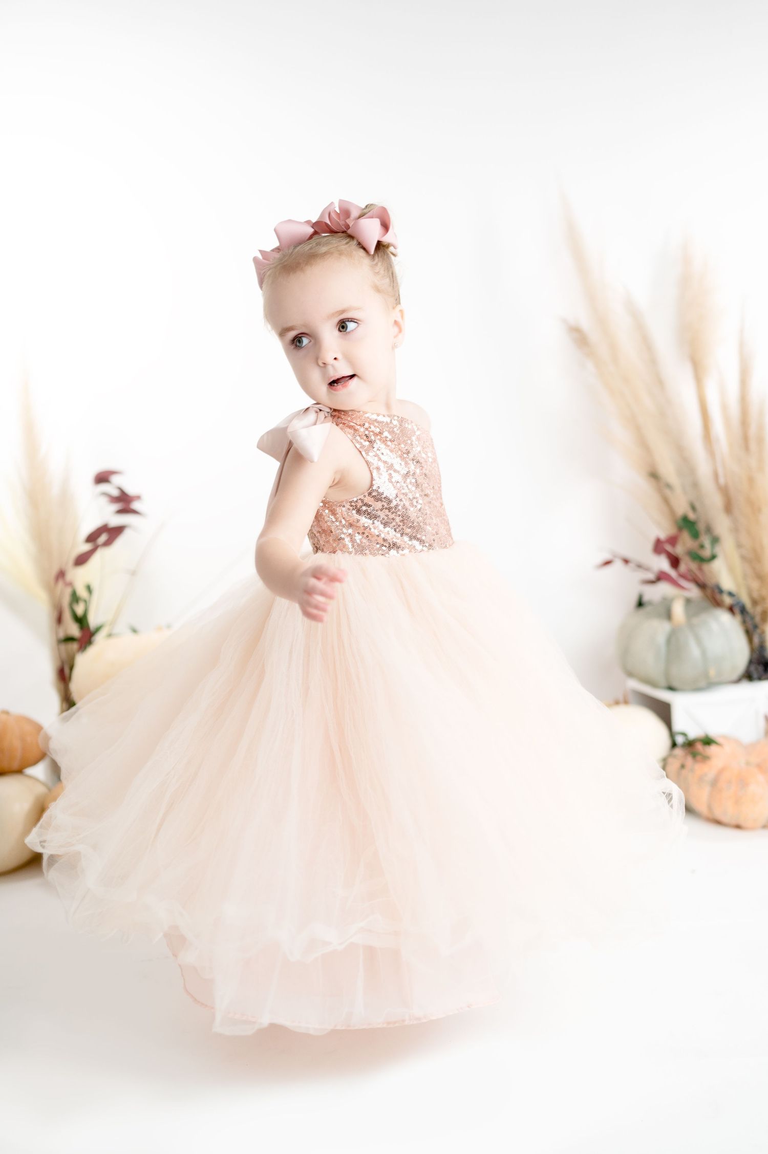 Small child in a blush pink tulle dress poses with pumpkins and dried flowers in a light studio setting.