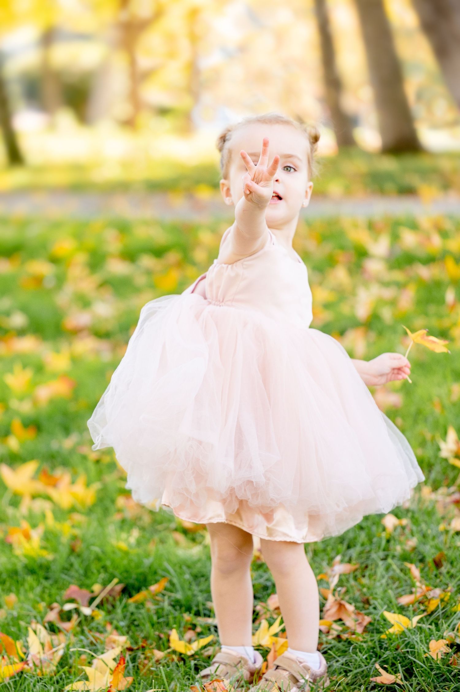 Child in flowing pink dress twirling among fallen autumn leaves in a sunlit park.