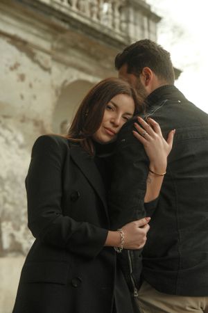 A romantic couple in dark coats embraces against a weathered stone wall backdrop.