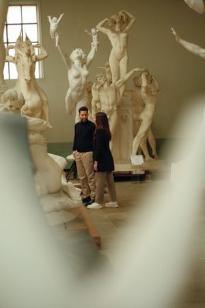 A couple stands in an art gallery surrounded by classical nude sculptures and statues against a beige wall.