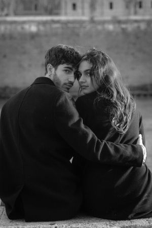 Couple sitting close together on stone wall in moody black and white portrait.