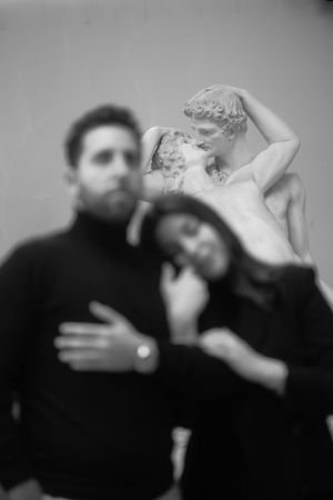 A moody black and white portrait of a couple embracing near a classical sculpture.