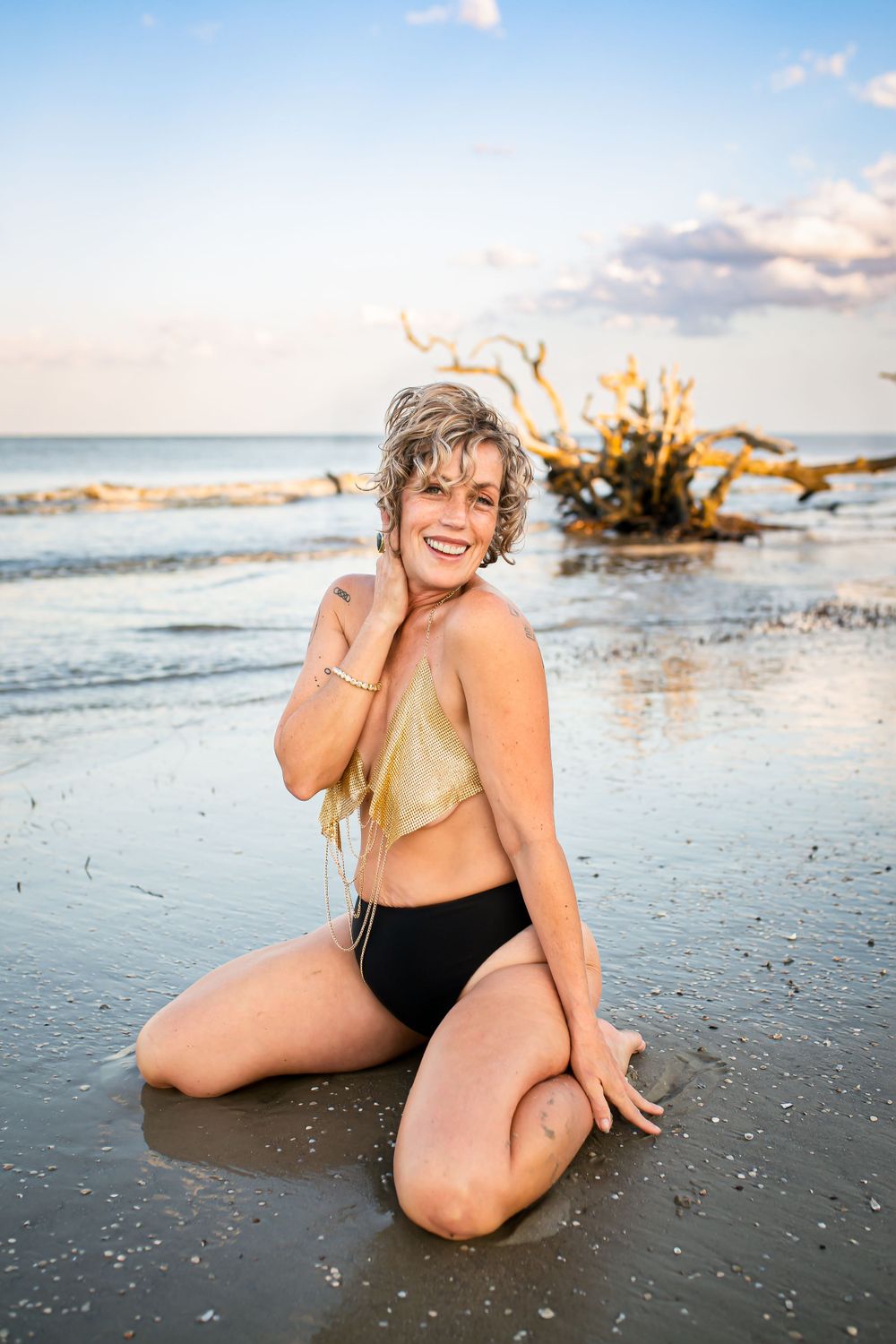 Person posing on wet beach sand at sunset with driftwood and ocean in background.