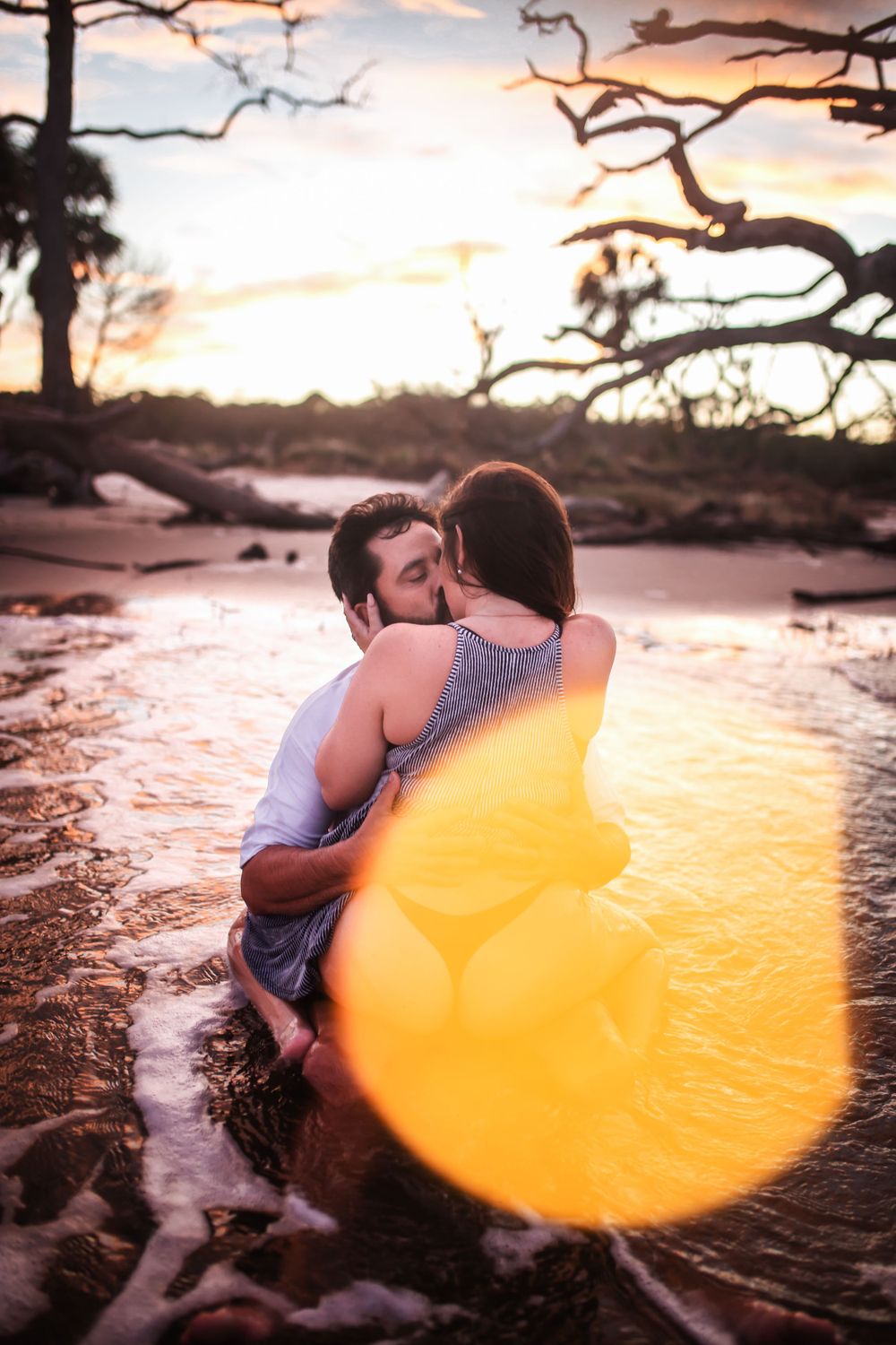 Romantic couple in water at sunset with golden lens flare and driftwood trees.