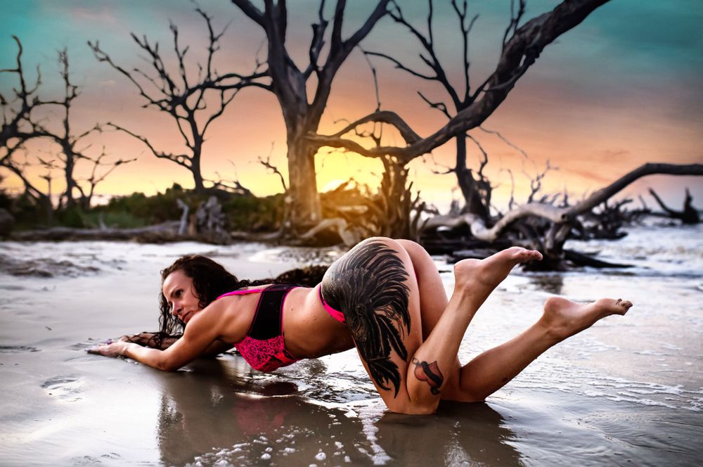 Two people posing on wet beach sand at sunset with driftwood trees in background.