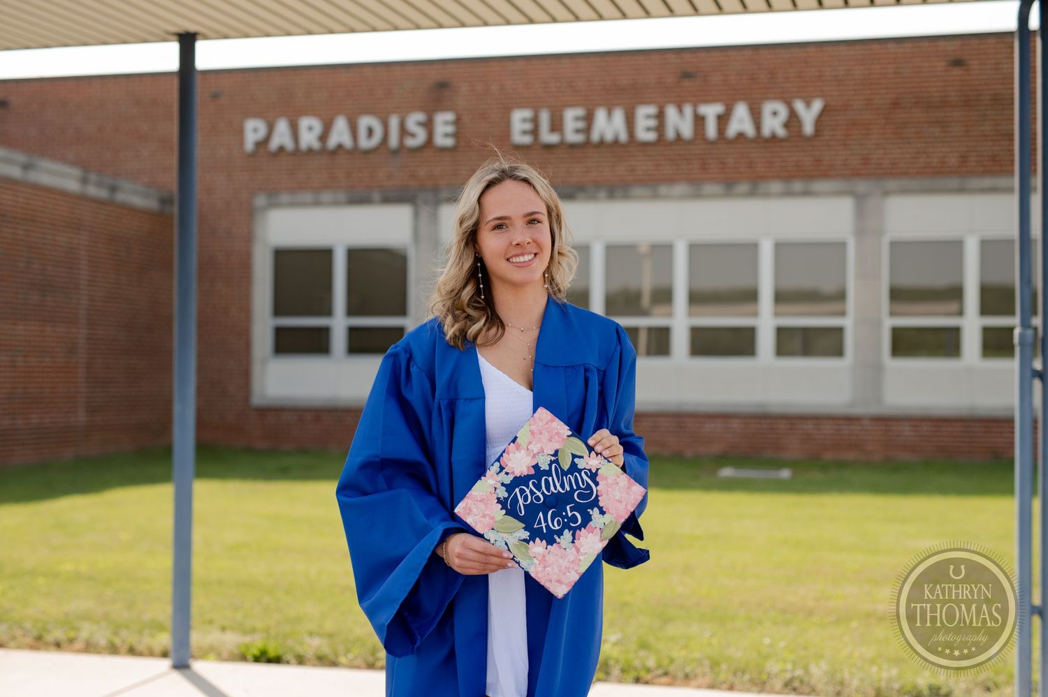 Graduate holds diploma while wearing blue graduation gown outside brick elementary school.